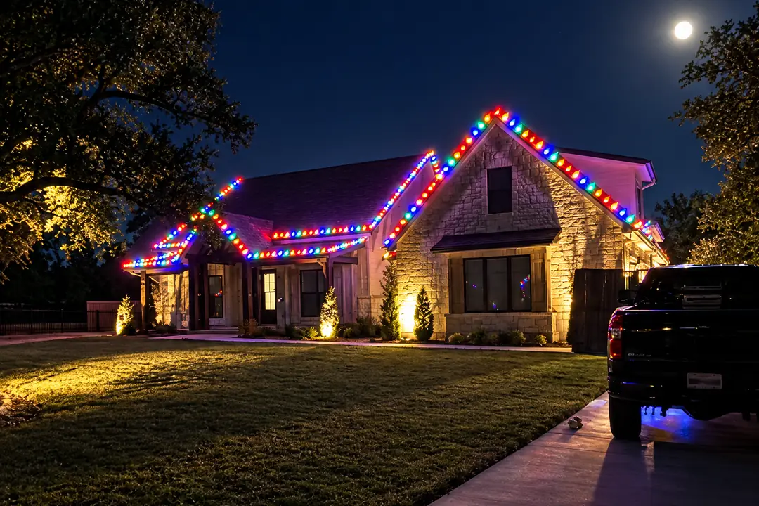 A stone farmhouse at dusk with multicolour lights along the gables, a full moon above the roof and a pickup in the driveway.