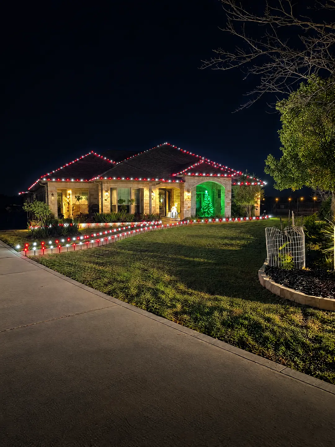 A stone and brick home at night with red and white lights along its rooflines, a green-lit arched entrance with a small lit tree inside, and red and white lights on stakes along the walkway and lawn.