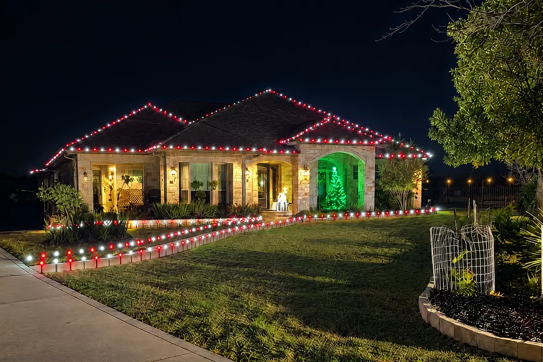 A stone and brick home at night with red and white lights along its rooflines, a green-lit arched entrance with a small lit tree inside, and red and white lights on stakes along the walkway and lawn.